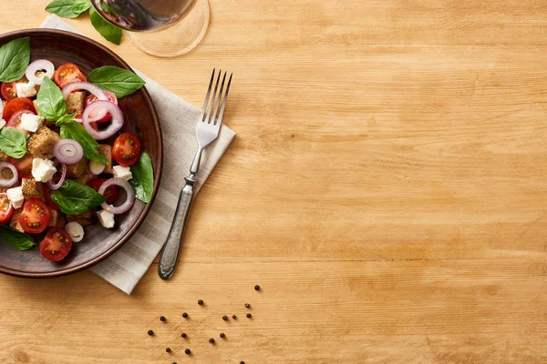 Top view of delicious Italian vegetable salad panzanella served on plate on wooden table near fork on napkin and red wine — Stock Photo