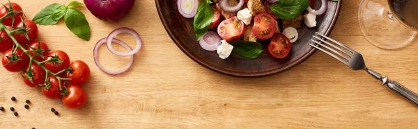 Vista dall'alto della deliziosa insalata di verdure italiana panzanella servita su piatto con forchetta su tavola di legno vicino a ingredienti freschi e vino rosso, colpo panoramico — Foto stock