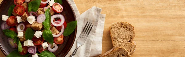 Vista dall'alto della deliziosa insalata di verdure italiana panzanella servita sul piatto sul tavolo di legno vicino a pane e forchetta sul tovagliolo, colpo panoramico — Foto stock