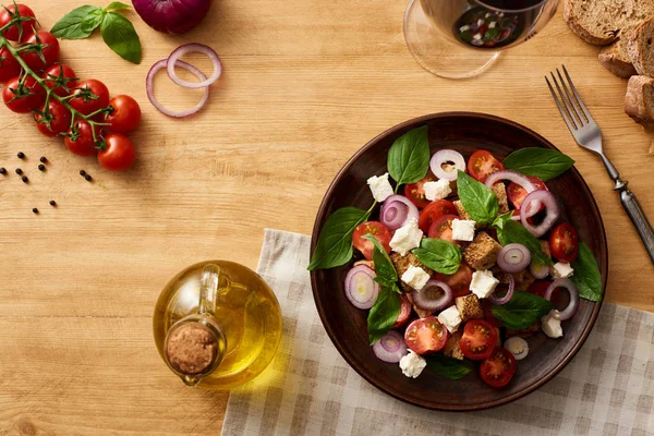 Сверху вид на вкусный итальянский овощной салат panzanella подается на тарелке на деревянном столе рядом со свежими ингредиентами и красным вином — стоковое фото
