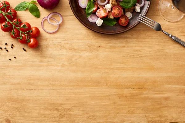 Vista superior da deliciosa salada de legumes italiana panzanella servida em prato em mesa de madeira perto de ingredientes frescos e vinho tinto — Fotografia de Stock