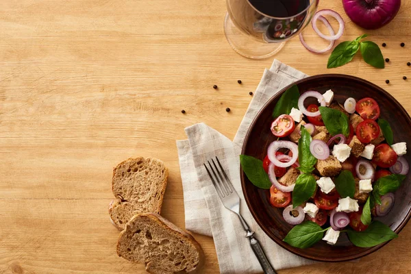 Vista superior da deliciosa salada de legumes italiana panzanella servida em prato em mesa de madeira perto de ingredientes frescos, garfo, pão e vinho tinto — Fotografia de Stock