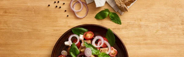 Blick von oben auf köstlichen italienischen Gemüsesalat Panzanella serviert auf Teller auf Holztisch in der Nähe von frischen Zutaten, Panoramaaufnahme — Stockfoto