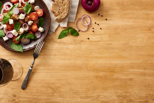Vista superior de la deliciosa ensalada de verduras italiana panzanella servida en plato con tenedor en mesa de madera cerca de ingredientes frescos en servilleta y vino tinto - foto de stock