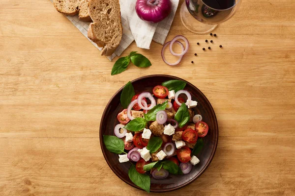 Vue du dessus de la délicieuse salade de légumes italienne panzanella servi sur une assiette sur une table en bois près d'ingrédients frais sur une serviette et du vin rouge — Photo de stock