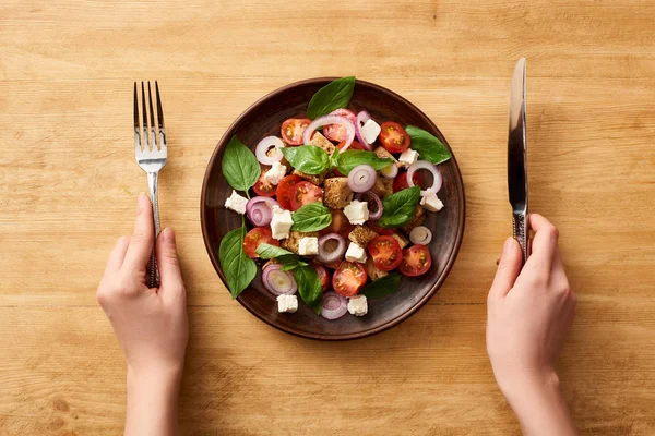 Ausgeschnittene Ansicht einer Frau, die Panzanella-Salat mit Messer und Gabel am Holztisch isst — Stockfoto