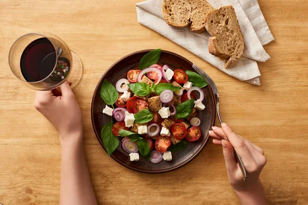 Vista recortada de la mujer comiendo ensalada de panzanella y beber vino tinto en la mesa de madera - foto de stock