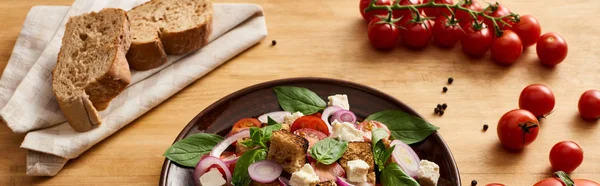 Deliciosa salada de legumes italiana panzanella servido em prato na mesa de madeira perto de tomates e pão, tiro panorâmico — Fotografia de Stock