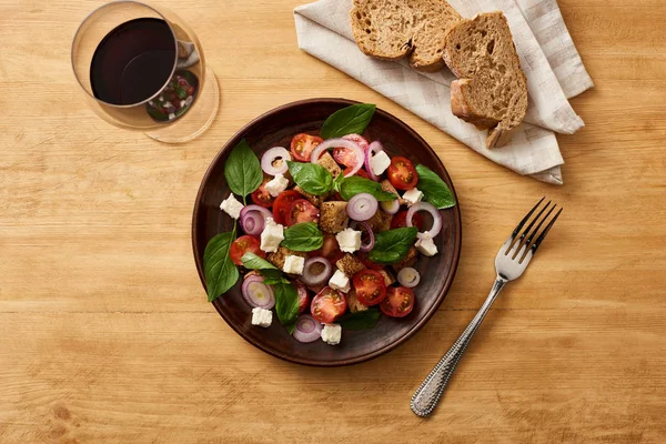Top view of delicious Italian vegetable salad panzanella served on plate on wooden table near fresh bread and red wine — Stock Photo