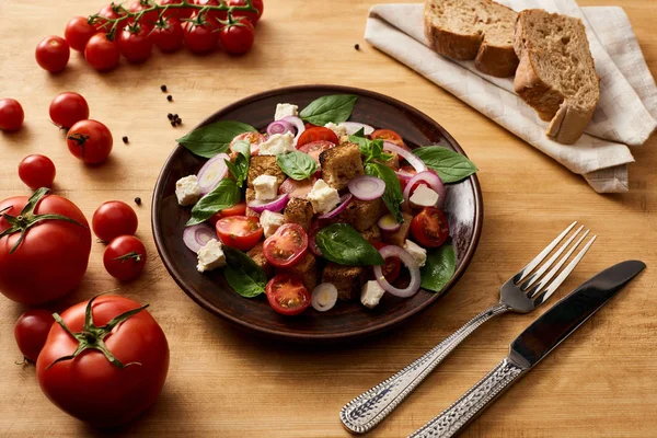Смачна італійська салатна панцанела подається на тарілці на дерев'яному столі біля свіжих помідорів, хліба, виделки та ножа. — стокове фото