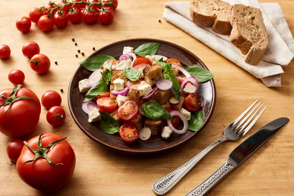 Deliziosa insalata di verdure italiana panzanella servita sul piatto sul tavolo di legno vicino a pomodori freschi, pane, forchetta e coltello — Foto stock