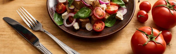 Deliziosa insalata di verdure italiana panzanella servita sul piatto sul tavolo di legno vicino a pomodori freschi, forchetta e coltello, colpo panoramico — Foto stock