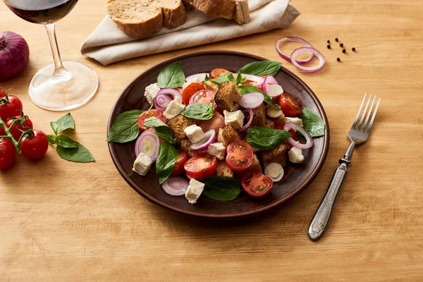 Вкусный итальянский овощной салат panzanella подается на тарелке на деревянном столе рядом со свежими ингредиентами, вилка, хлеб и красное вино — стоковое фото