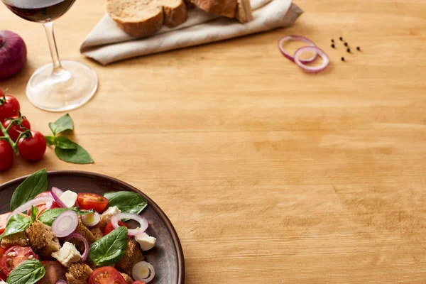 Смачна італійська салатна панцанела подається на тарілці на дерев'яному столі біля свіжих інгредієнтів, хліба та червоного вина. — стокове фото