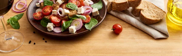 Deliziosa insalata di verdure italiana panzanella servita su piatto su tavola di legno vicino a ingredienti freschi, pane e vino rosso, colpo panoramico — Foto stock