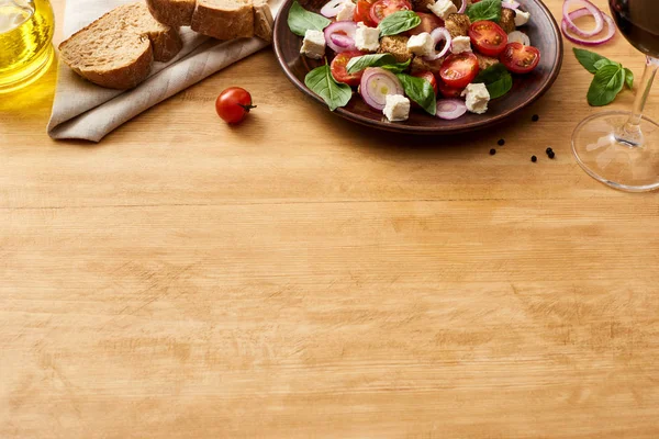 Вкусный итальянский овощной салат panzanella подается на тарелке на деревянном столе рядом со свежими ингредиентами, хлеб и красное вино — стоковое фото
