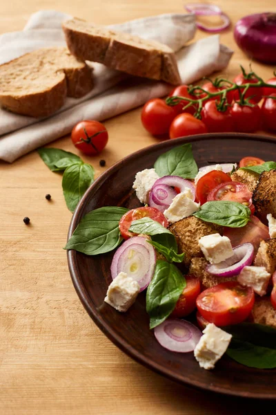 Deliciosa ensalada de verduras italiana panzanella servido en el plato en la mesa de madera cerca de ingredientes frescos y pan - foto de stock