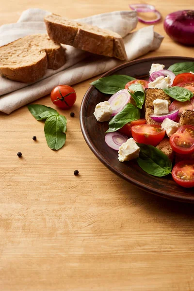 Deliciosa salada de legumes italiana panzanella servida em prato em mesa de madeira perto de ingredientes frescos e pão — Fotografia de Stock