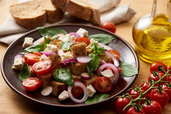 Селективный фокус вкусного итальянского овощного салата panzanella подается на тарелке на деревянном столе рядом со свежими помидорами, оливковым маслом и хлебом — стоковое фото