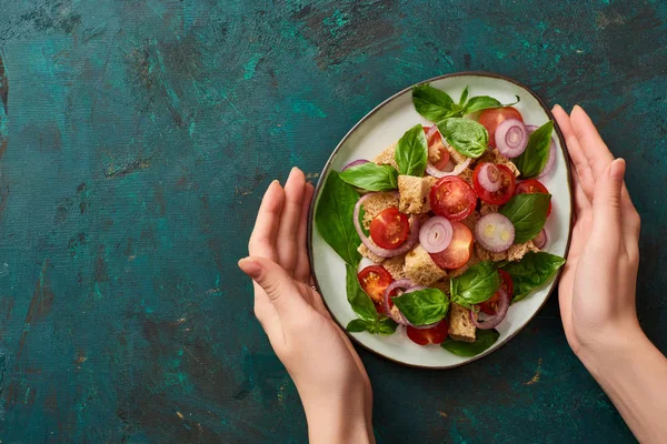 Cropped view of woman holding plate with delicious Italian vegetable salad panzanella on textured green surface — Stock Photo