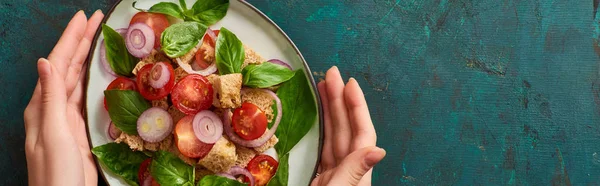 Cropped view of woman holding plate with delicious Italian vegetable salad panzanella on textured green surface, panoramic shot — Stock Photo