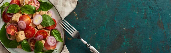 Top view of delicious Italian vegetable salad panzanella served on plate on textured green surface with napkin and fork, panoramic shot — Stock Photo