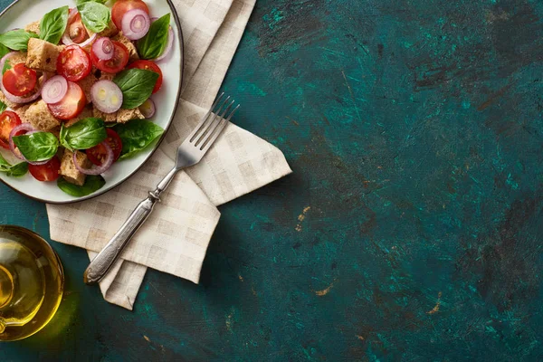 Верхний вид на вкусный итальянский овощной салат panzanella подается на тарелке на текстурированной зеленой поверхности с салфеткой и вилкой — стоковое фото