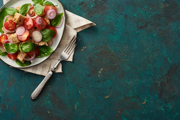Vue du dessus de la délicieuse salade de légumes italienne panzanella servie sur assiette sur une surface verte texturée avec serviette et fourchette — Photo de stock