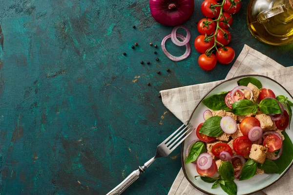 Vista superior de la deliciosa ensalada de verduras italiana panzanella servido en el plato en la superficie verde con textura con ingredientes, servilleta y tenedor - foto de stock