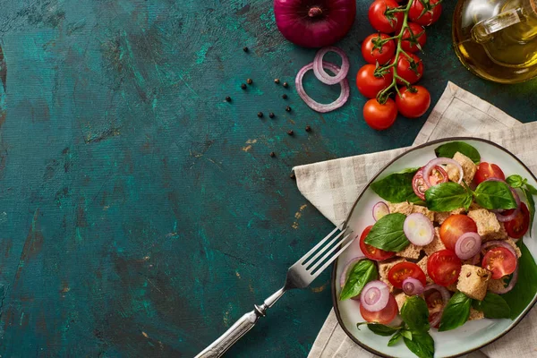 Top view of delicious Italian vegetable salad panzanella served on plate on textured green surface with ingredients, napkin and fork — Stock Photo