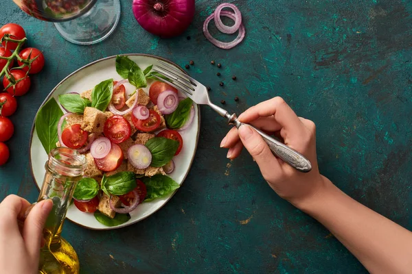 Vista recortada de la mujer añadiendo aceite de oliva a la deliciosa ensalada de verduras italiana panzanella en la superficie verde texturizada con ingredientes — Stock Photo