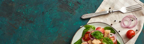 Сверху вид на вкусный итальянский овощной салат panzanella подается на тарелке на текстурированной зеленой поверхности с ингредиентами, салфеткой и вилкой, панорамный снимок — стоковое фото