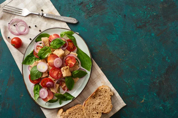 Верхній вигляд смачного італійського салату панцанелла подається на тарілці на текстурованій зеленій поверхні з хлібом, інгредієнтами, серветкою і виделкою. — стокове фото