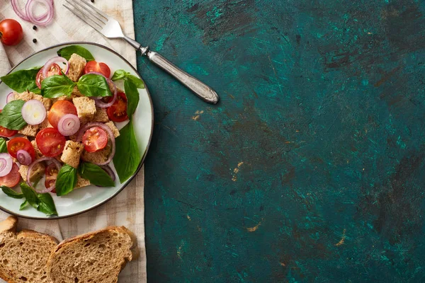 Vista superior da deliciosa salada de legumes italiana panzanella servida em prato na superfície verde texturizada com pão, ingredientes, guardanapo e garfo — Fotografia de Stock