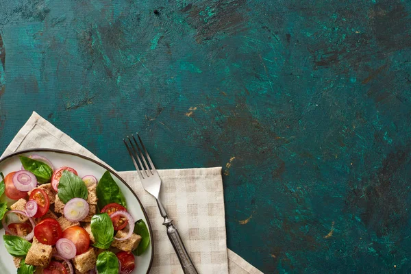 Top view of delicious Italian vegetable salad panzanella served on plate on textured green surface with napkin and fork — Stock Photo