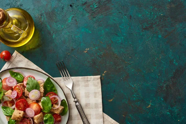 Vista dall'alto della deliziosa insalata di verdure italiana panzanella servita su piatto su superficie verde strutturata con olio d'oliva, tovagliolo e forchetta — Foto stock