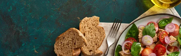 Top view of delicious Italian vegetable salad panzanella served on plate on textured green surface with olive oil, bread, napkin and fork, panoramic shot — Stock Photo