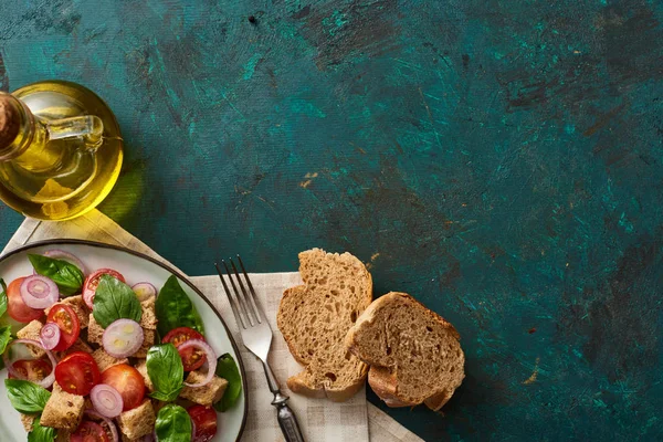 Vista superior da deliciosa salada de legumes italiana panzanella servida em prato na superfície verde texturizada com azeite, pão, guardanapo e garfo — Fotografia de Stock