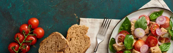 Top view of delicious Italian vegetable salad panzanella served on plate on textured green surface with bread, tomatoes, napkin and fork, panoramic shot — Stock Photo