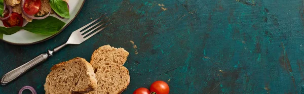 Top view of delicious Italian vegetable salad panzanella served on plate on textured green surface with bread and fork, panoramic shot — Stock Photo