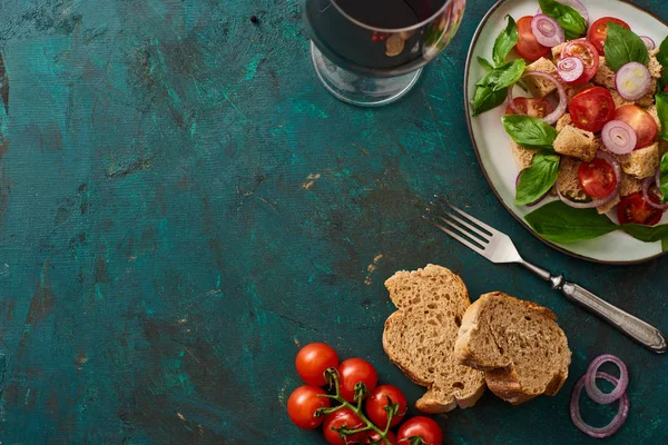 Vista superior da deliciosa salada de legumes italiana panzanella servida em prato na superfície verde texturizada com tomate, pão, vinho tinto e garfo — Fotografia de Stock