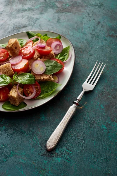 Вкусный итальянский овощной салат panzanella подается на тарелке на текстурированной зеленой поверхности с вилкой — стоковое фото