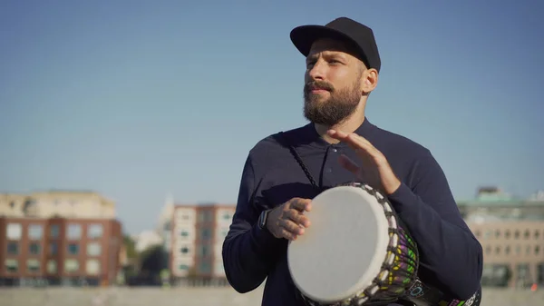 A musician stands on the street and holds a drum tomtom in his hands. A street musician plays tom-tom on a drum.