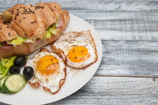 Fehér tál sült tojás és Croissant szendvics paradicsomos, uborka, olajbogyó. Reggeli. Szürke fa háttér. Rusztikus stílusú másolás beillesztés. — Stock Fotó