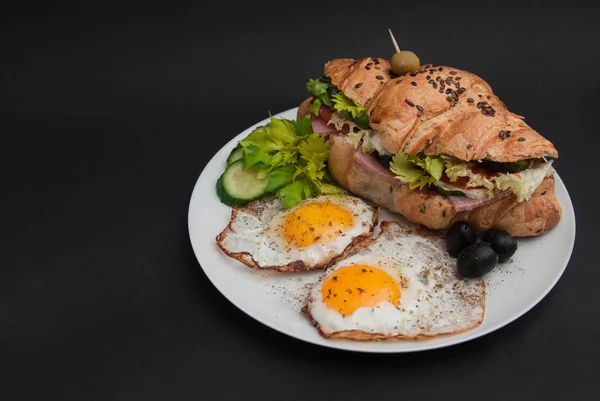 Snídaňový talíř. Smažená vejce s croissantem sendvič v bílé desky s čerstvou zeleninou černé pozadí s Copy prostor. — Stock fotografie