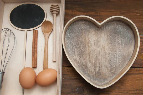 Plat leggen van voedsel proces achtergrond. Rauwe eieren met houten spooon en bakken van hulpmiddelen. Bak of voedsel achtergrond. Donkere rustieke houten achtergrond en Heart Shape Box, met kopie ruimte. Platte Lay. — Stockfoto