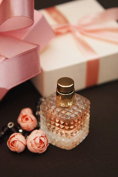 Frauen rosa Parfüm in schönen Flasche und künstliche Blumen Armband auf braunem Hintergrund mit Geschenkboxen auf dem Hintergrund. vertikales Bild. — Stockfoto