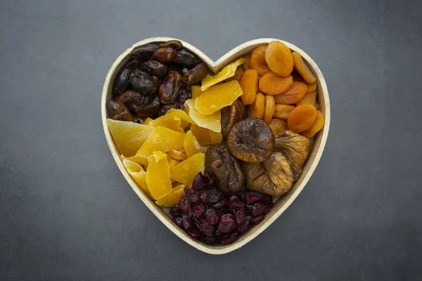 Dried fruits mix, in wooden heart shape box isolated on dark background. Top view of various dried fruits figs, apricots, mango, cranberries.