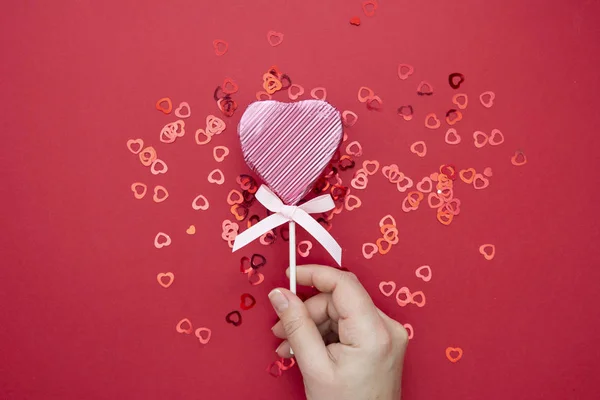 Día de San Valentín. Mano femenina sosteniendo una piruleta rosa en forma de corazón aislado sobre fondo rojo, con confeti espumoso . — Foto de Stock