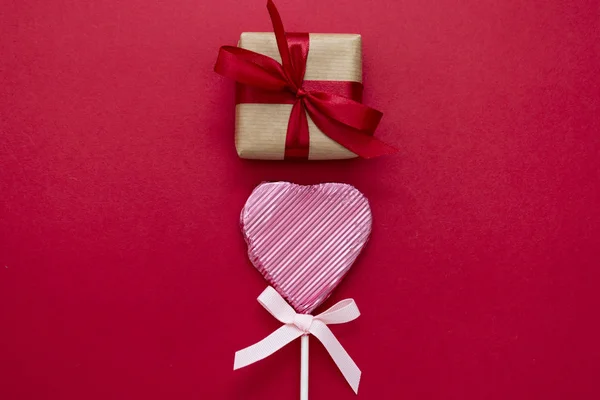 Amor, San Valentín se burlan, con piruleta en forma de corazón y caja de regalo, aislado sobre fondo rojo, espacio de copia . — Foto de Stock